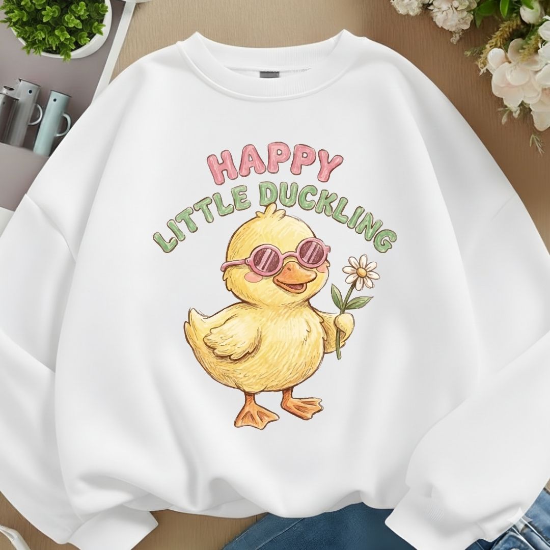 A white crewneck sweatshirt featuring a cartoon graphic of a yellow duckling wearing pink sunglasses and holding a daisy, with the text 'HAPPY LITTLE DUCKLING' above it. The sweatshirt is styled flat on a light wood background with decorative plants and flowers.