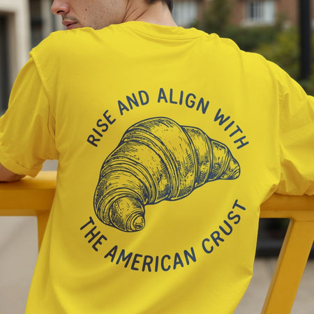 Rear view of a person wearing a bright yellow t-shirt printed with a large blue croissant sketch and bakery slogan across the back.