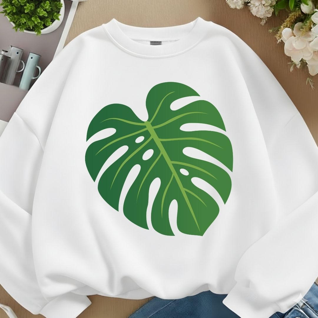 A white crewneck sweatshirt featuring a large green monstera leaf graphic on the front, styled in a flat-lay arrangement with a wooden background, plants, and flowers.