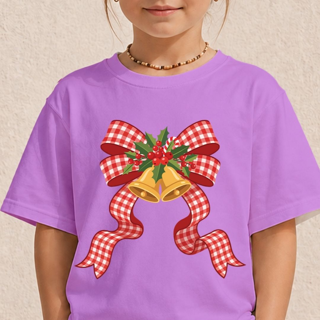 A child wearing a lavender purple t-shirt with a festive Christmas graphic of two gold bells, a red and white gingham bow, and green holly leaves on the front, set against a textured beige wall.