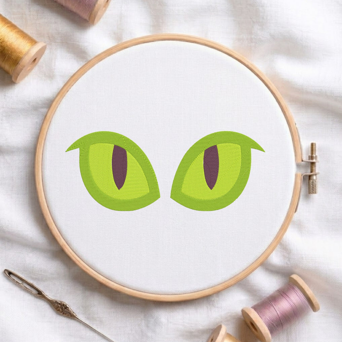 A wooden embroidery hoop holding white fabric with a simple green cat-eye design, surrounded by thread spools and a needle on a white cloth background.