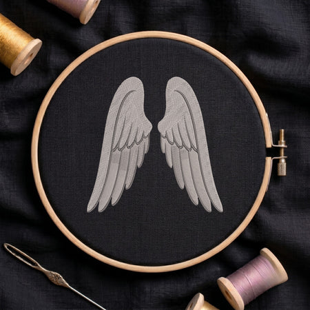 A wooden embroidery hoop holds black fabric with a stitched gray angel wings design in the center. Spools of thread, a needle, and dark fabric are arranged around it in a styled flat-lay.