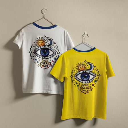 Two ringer t-shirts with a blue collar hanging on metal hangers against a beige wall. The left shirt is white with a large graphic on the back featuring a blue eye, sun, and moon with the text 'GOOD ENERGY ONLY'.
