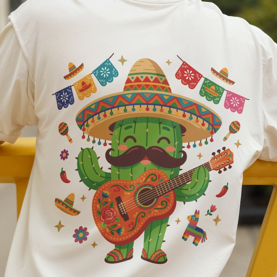 A white short-sleeve T-shirt featuring a colorful cartoon cactus wearing a large sombrero, black mustache, and playing a decorated guitar, surrounded by festive Mexican-themed accents like papel picado banners, maracas, chili peppers, and a piñata. The shirt is shown on a person, cropped from the neck down, with a yellow chair visible in the background.