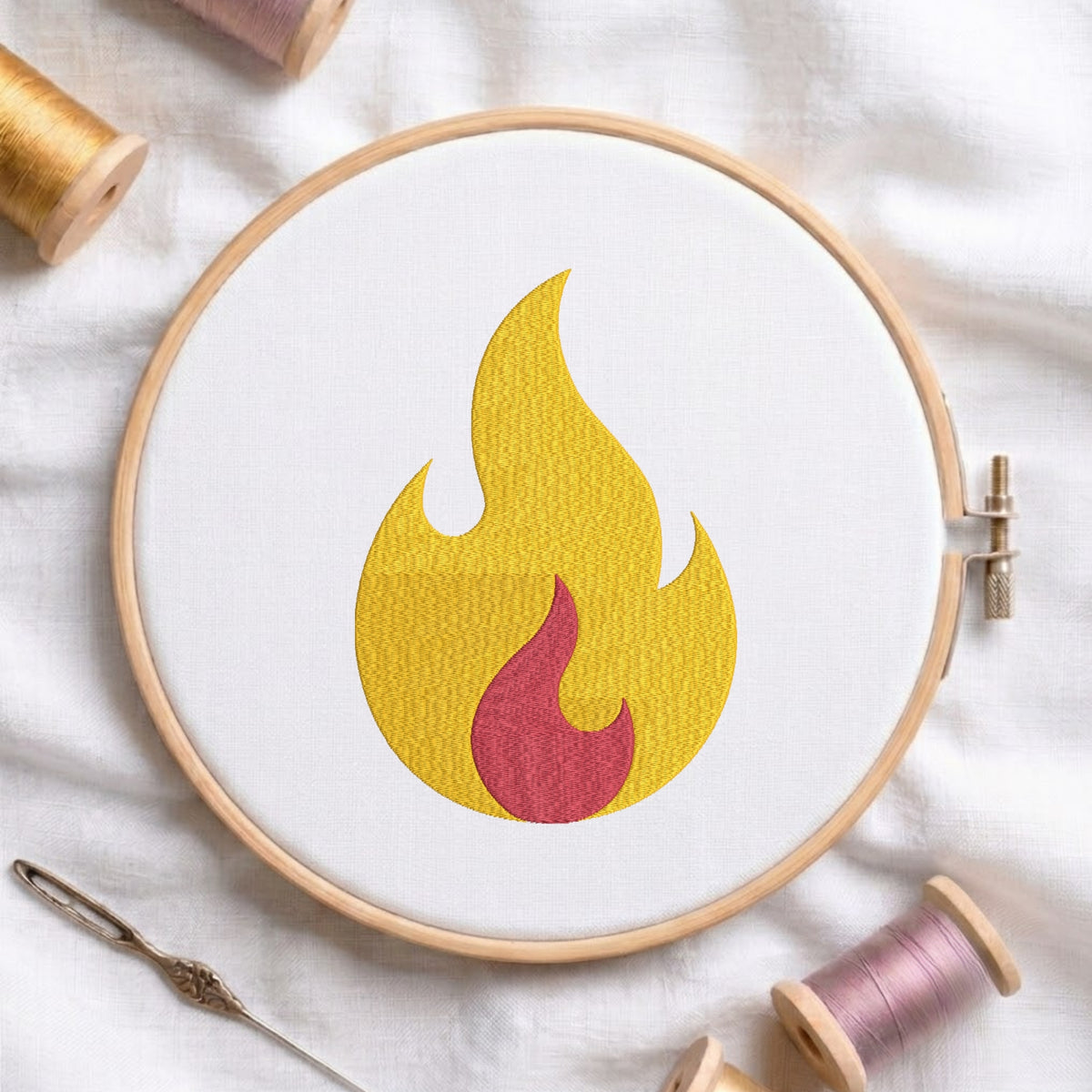 A circular embroidery hoop on white fabric displays a flame motif in yellow and red. Surrounding props include spools of thread, a needle, and a wooden spool, arranged on a soft white textile background.