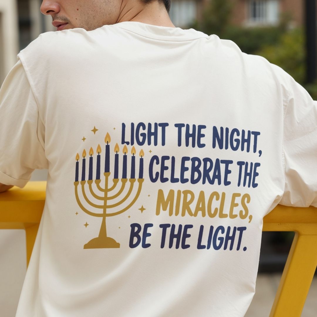 A man wearing a white t-shirt from behind, featuring a large graphic print with a gold menorah and the text 'LIGHT THE NIGHT, CELEBRATE THE MIRACLES, BE THE LIGHT.' in navy and gold lettering.