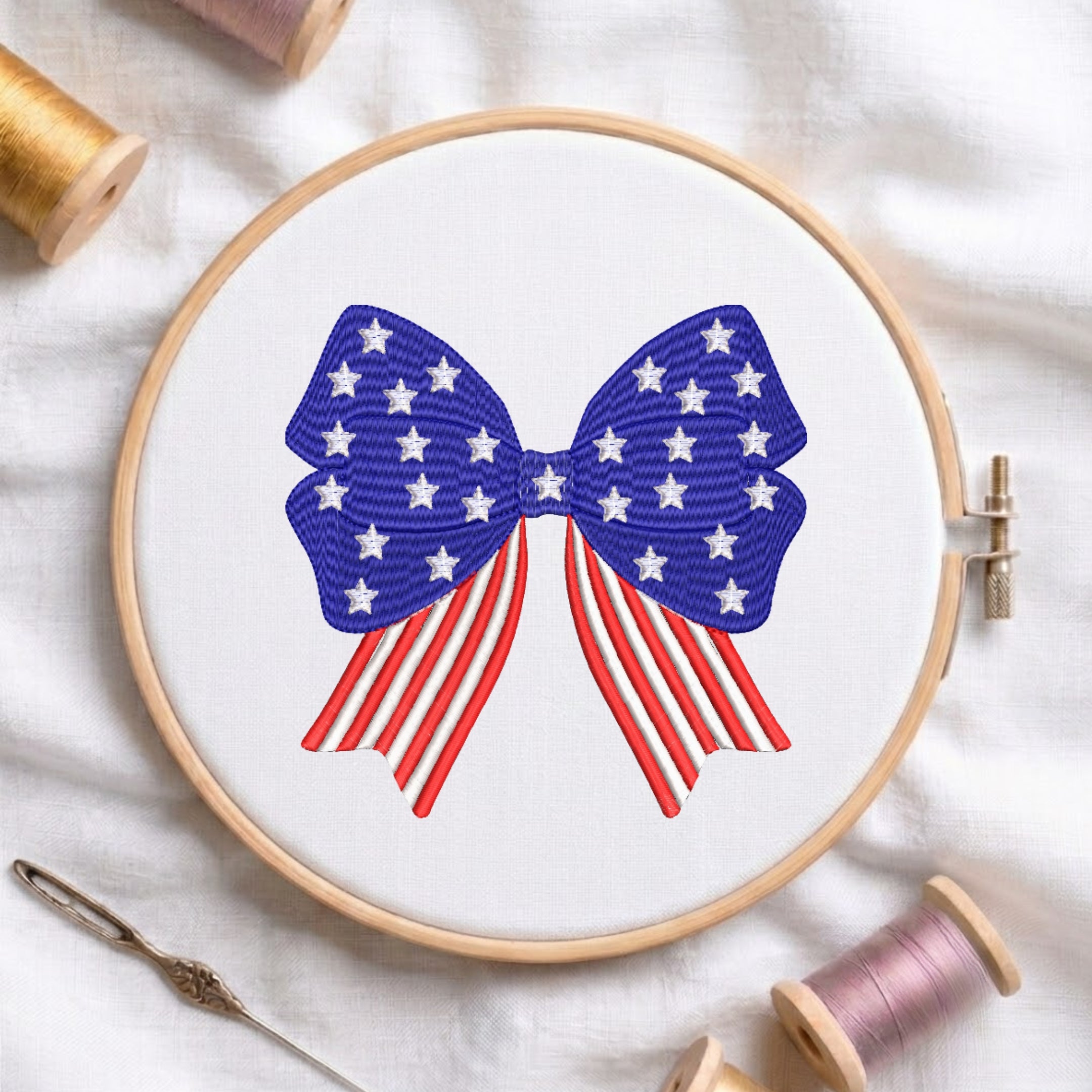 A wooden embroidery hoop on white fabric, featuring a bow design with white stars on a blue background and red-and-white stripes below, surrounded by spools of thread and a needle.