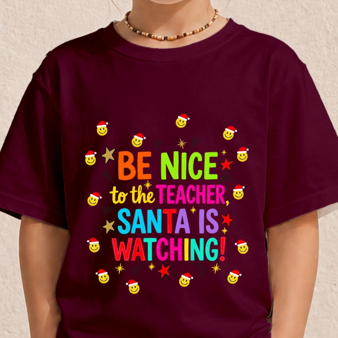A person wearing a dark burgundy t-shirt with a colorful Christmas-themed graphic that reads 'BE NICE to the TEACHER, SANTA IS WATCHING!' surrounded by smiling faces wearing Santa hats and gold stars.