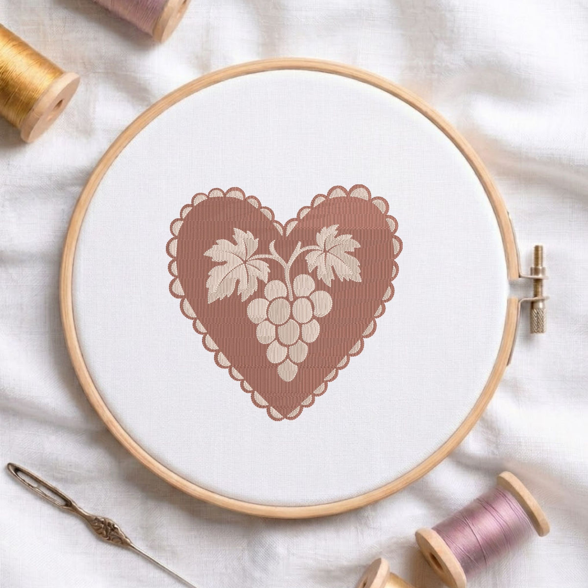 Vintage heart-shaped grape cluster machine embroidery design framed with a scalloped border, displayed in a wooden hoop on white fabric with thread spools and a needle.   