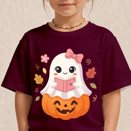 A child wearing a dark maroon short-sleeve t-shirt with a cute cartoon graphic of a white ghost reading a pink book on top of an orange jack-o'-lantern pumpkin, set against a textured beige wall.