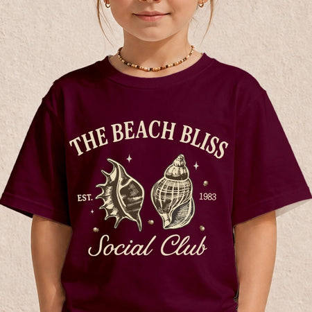 A young child wearing a burgundy t-shirt printed with a retro ocean design featuring two cream-colored sea shells and the text The Beach Bliss Social Club Est. 1983.