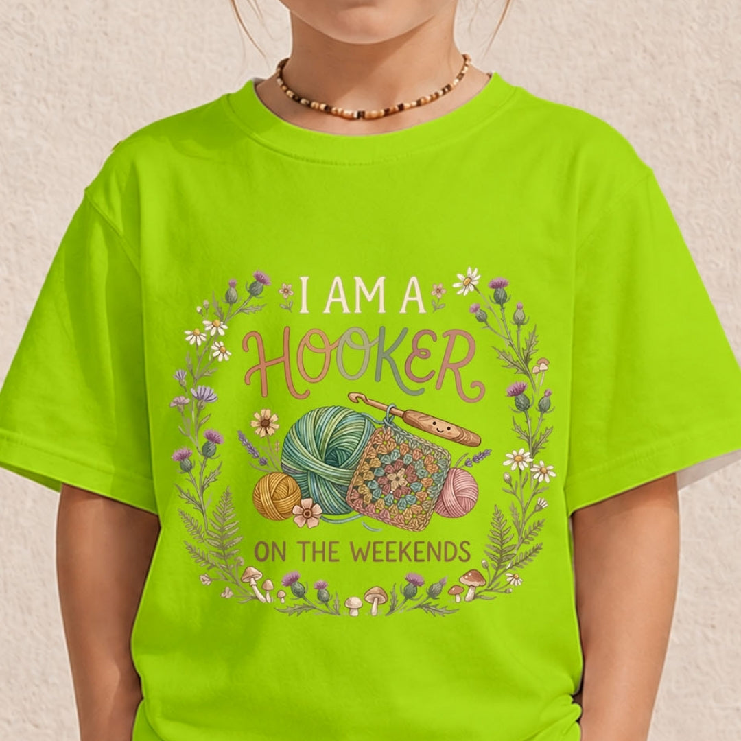 A child wearing a bright lime green t-shirt with a colorful graphic that reads 'I AM A HOOKER ON THE WEEKENDS' surrounded by yarn, crochet hooks, flowers, and mushrooms.