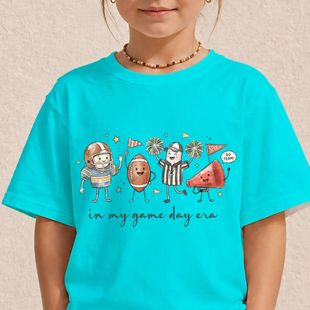 A child wearing a bright turquoise short-sleeve T-shirt with a colorful cartoon graphic of sports figures and the text 'in my game day era' on the front, photographed against a light textured wall.