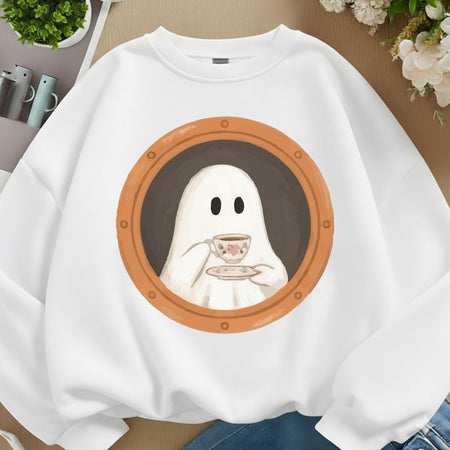 A white crewneck sweatshirt featuring a graphic print of a cute cartoon ghost peeking over a porthole window and holding a teacup. The sweatshirt is displayed in a styled flat-lay setting with a wooden surface, blue jeans, and small plants.
