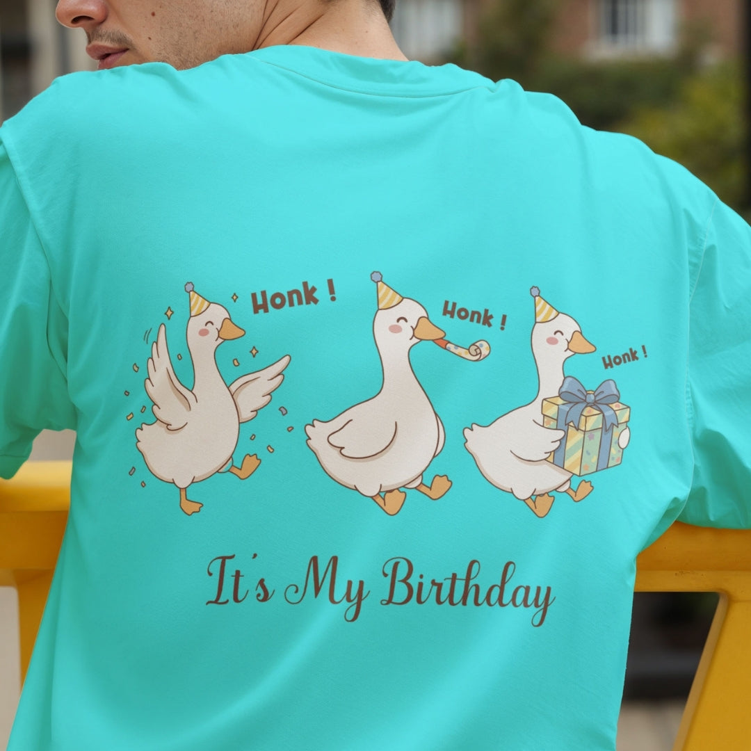 Back view of a person wearing a bright turquoise t-shirt showcasing a funny graphic of three partying geese wearing party hats, holding a present and a party blower, with the text "Honk! Honk! Honk! It's My Birthday".
