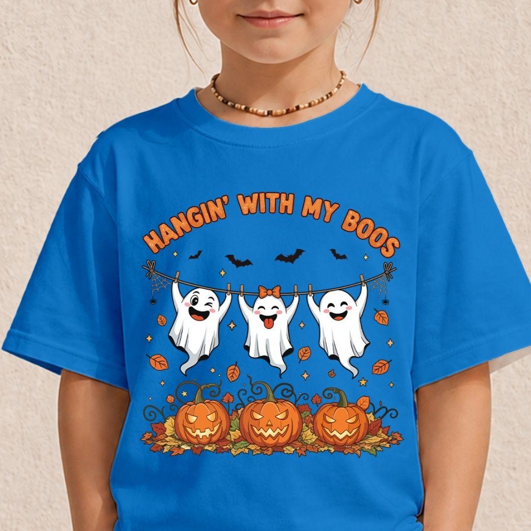 A young girl wearing a bright blue t-shirt printed with three cheerful ghosts hanging on a line above carved Halloween pumpkins and the phrase "HANGIN' WITH MY BOOS."