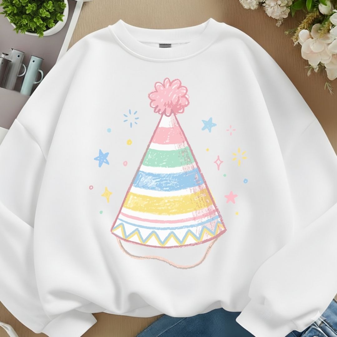 A white crewneck sweatshirt featuring a colorful pastel party hat graphic on the front, styled in a flat-lay arrangement with blue jeans, flowers, and small plants.