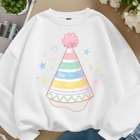 A white crewneck sweatshirt featuring a colorful pastel party hat graphic on the front, styled in a flat-lay arrangement with blue jeans, flowers, and small plants.