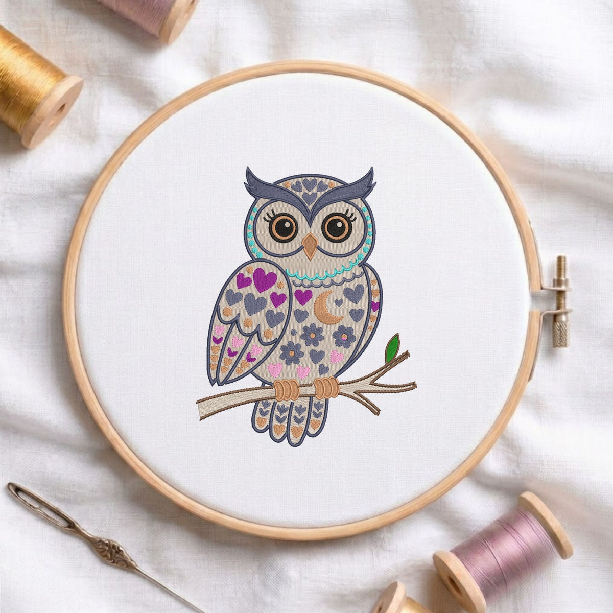 Detailed folk art owl machine embroidery design displayed inside a round wooden hoop on white fabric with thread spools and a needle.
