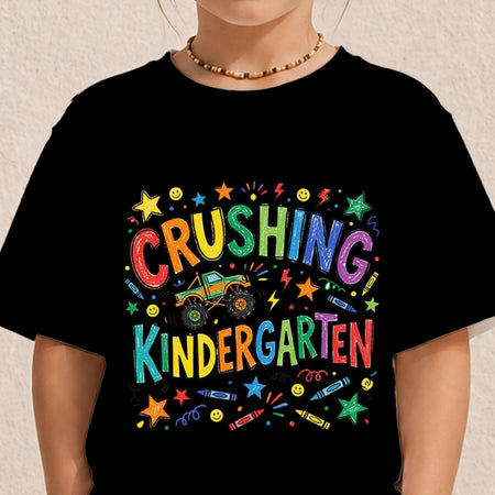 A black short-sleeve graphic T-shirt worn by a child, featuring the colorful text 'CRUSHING KINDERGARTEN' with a monster truck and stars.