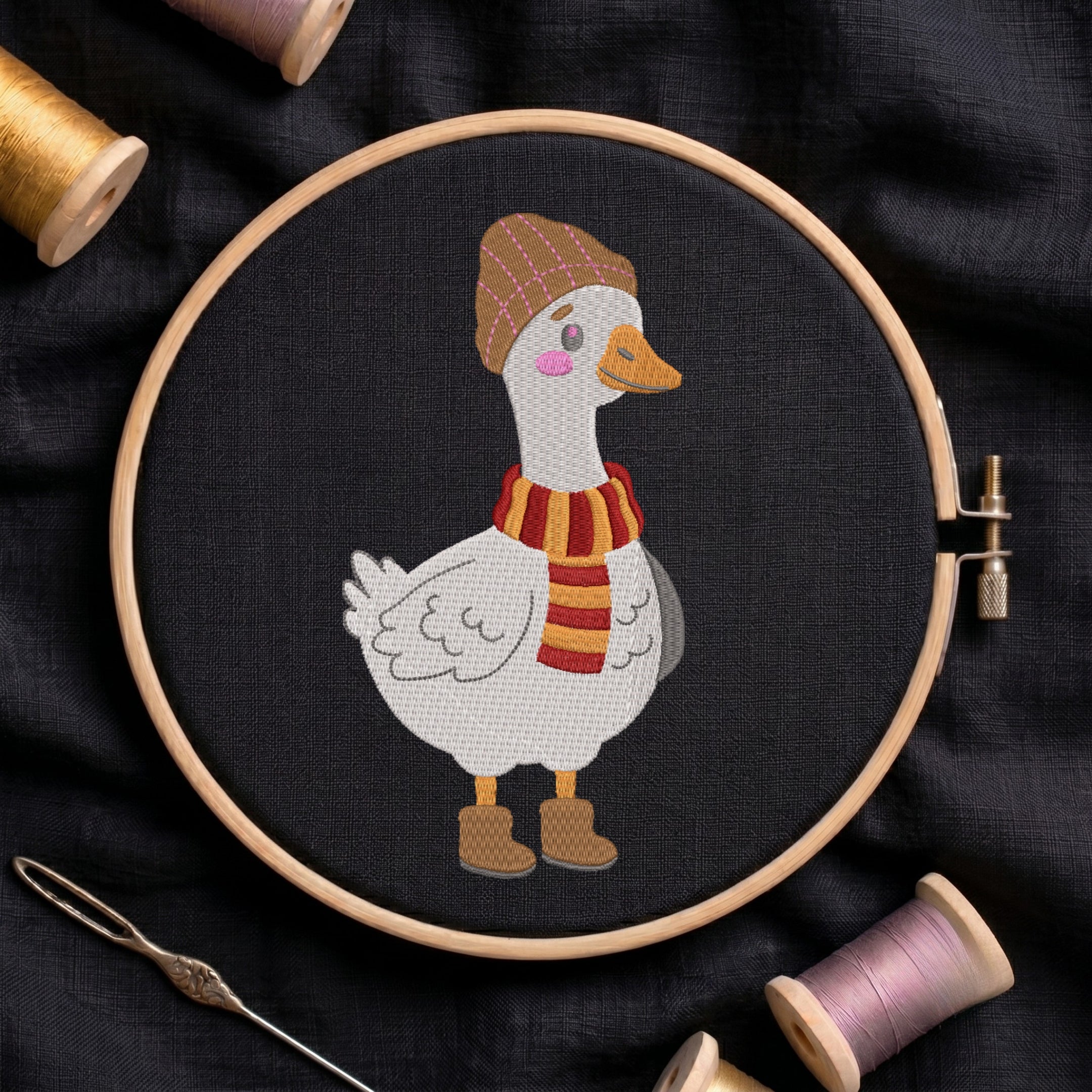A circular embroidery hoop holds a stitched white goose wearing a brown hat, orange beak, pink cheeks, and a red-and-orange striped scarf, with brown boots, on a dark fabric background. Spools of thread and a needle are arranged around it.