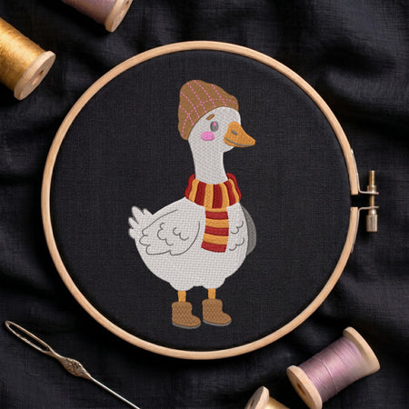 A circular embroidery hoop holds a stitched white goose wearing a brown hat, orange beak, pink cheeks, and a red-and-orange striped scarf, with brown boots, on a dark fabric background. Spools of thread and a needle are arranged around it.