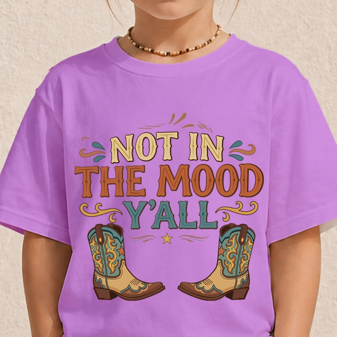 A child wearing a lavender purple graphic t-shirt with the text 'NOT IN THE MOOD Y'ALL' and cowboy boots printed on the front, set against a neutral textured wall.