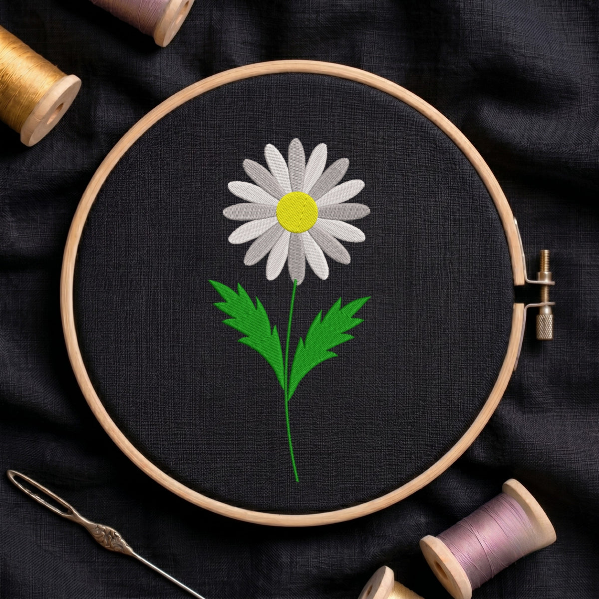 A wooden embroidery hoop holds black fabric with a stitched daisy flower design in white, yellow, and green. The hoop is photographed from above on a dark fabric background, with spools of thread and a needle partly visible around it.