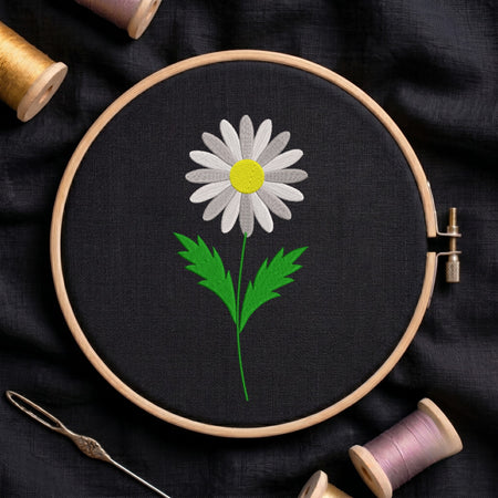 A wooden embroidery hoop holds black fabric with a stitched daisy flower design in white, yellow, and green. The hoop is photographed from above on a dark fabric background, with spools of thread and a needle partly visible around it.