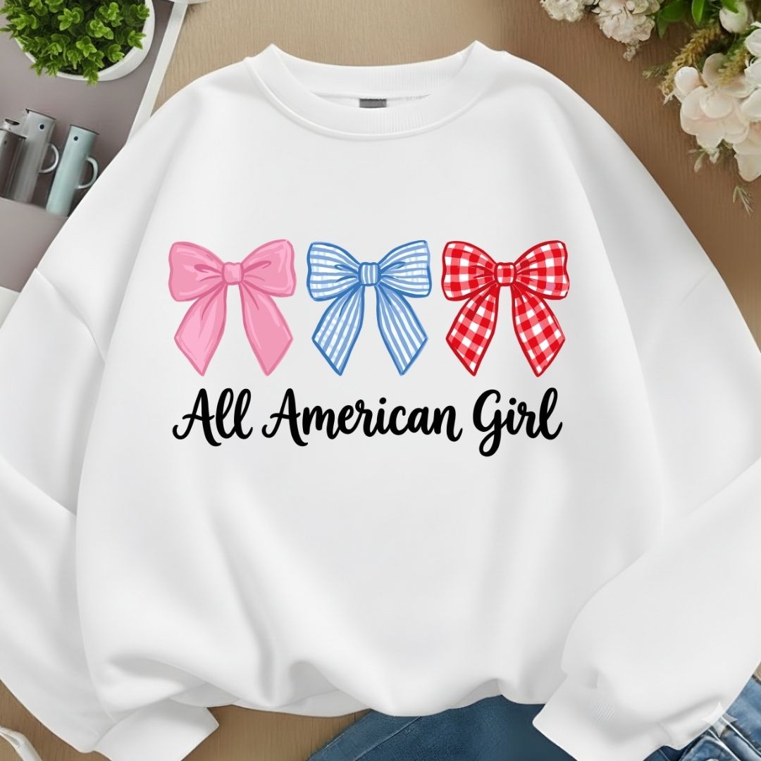 A white crewneck sweatshirt laid flat on a beige surface, featuring three decorative bows in pink, blue, and red gingham above the black script text 'All American Girl'.