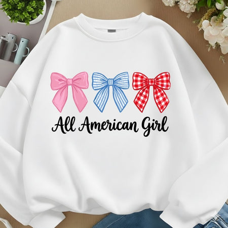 A white crewneck sweatshirt laid flat on a beige surface, featuring three decorative bows in pink, blue, and red gingham above the black script text 'All American Girl'.