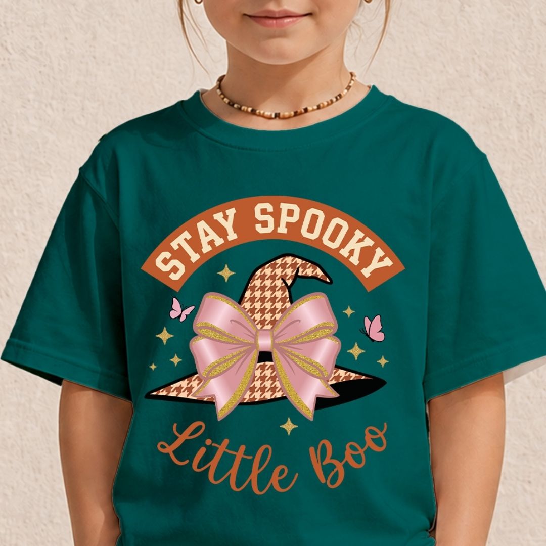 A child wearing a dark teal green graphic t-shirt with a 'Stay Spooky Little Boo' design featuring a witch hat and pink bow, against a textured beige wall.