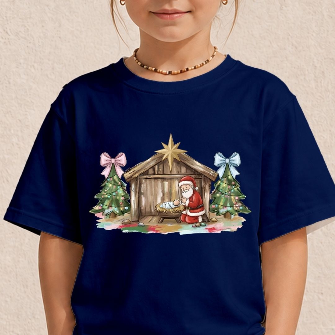 A child wearing a navy blue short-sleeve t-shirt with a colorful Christmas nativity scene graphic on the chest, featuring Santa Claus, a stable, and decorated trees.