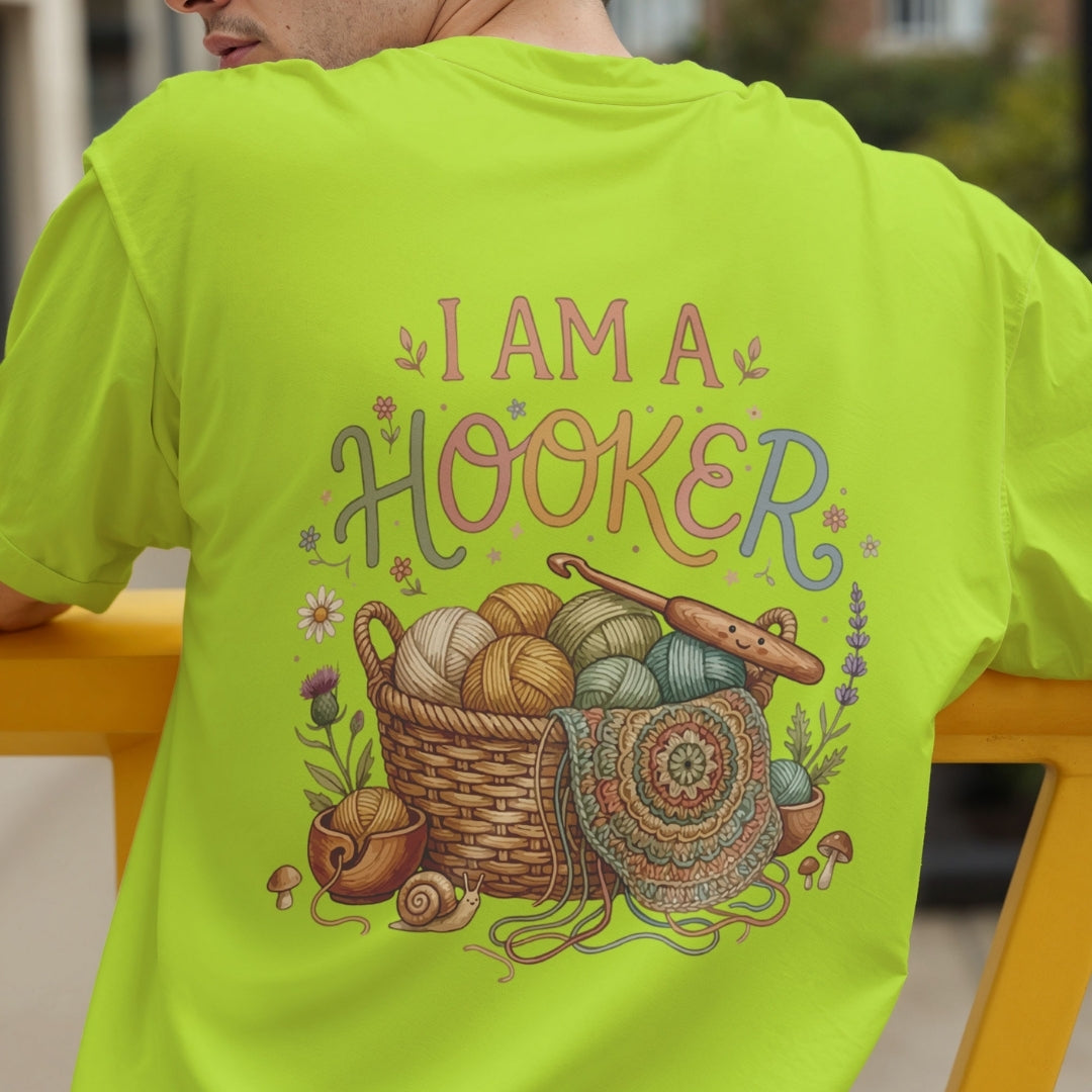 A person wearing a bright lime green t-shirt, viewed from the back. The shirt features a large, colorful graphic print with the text 'I AM A HOOKER' above a basket filled with colorful yarn balls, a crochet hook, and knitting needles.