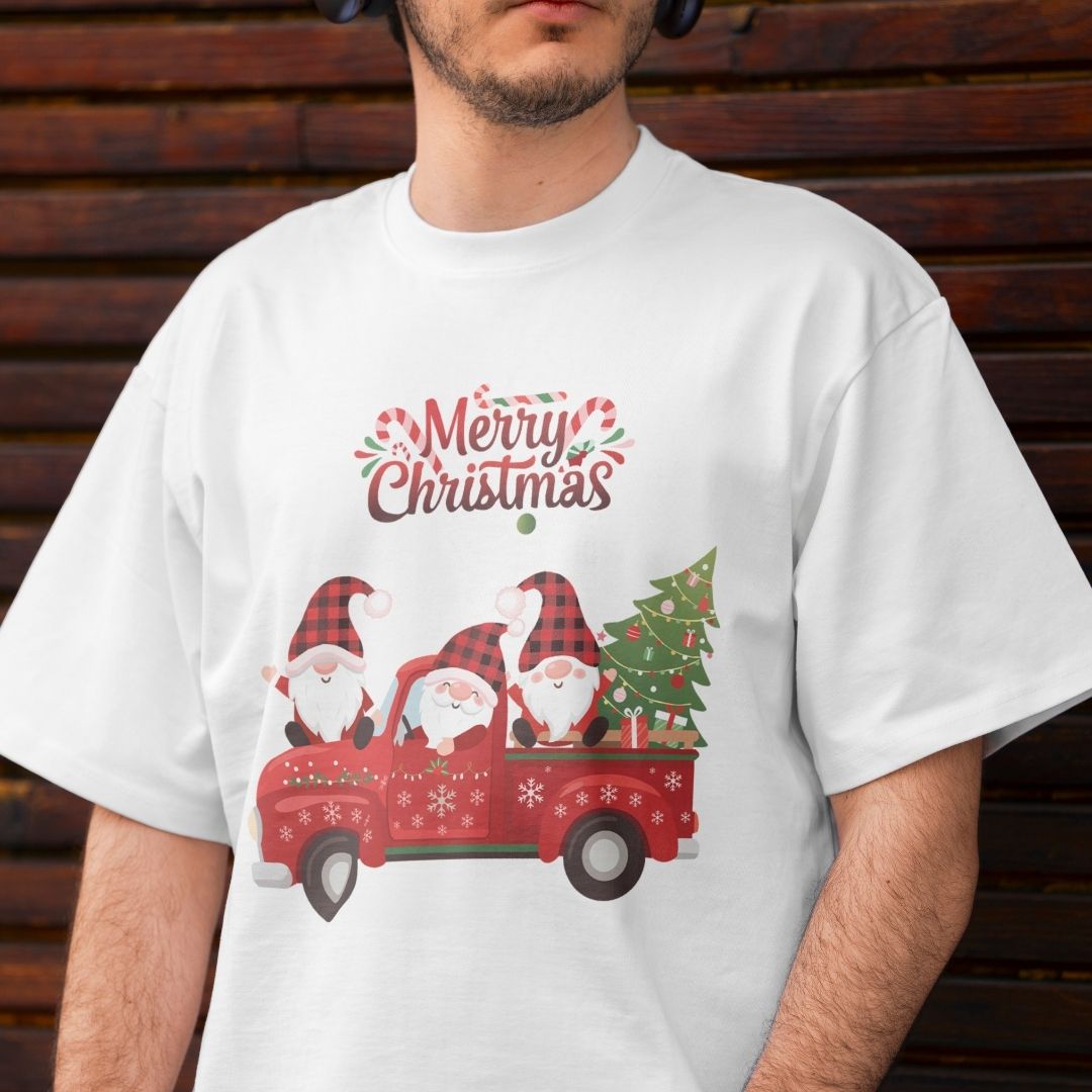 A man wearing a white crewneck t-shirt with a festive Christmas graphic. The graphic features three cartoon gnomes in a red pickup truck carrying a Christmas tree, with 'Merry Christmas' written above in a decorative red and green font.