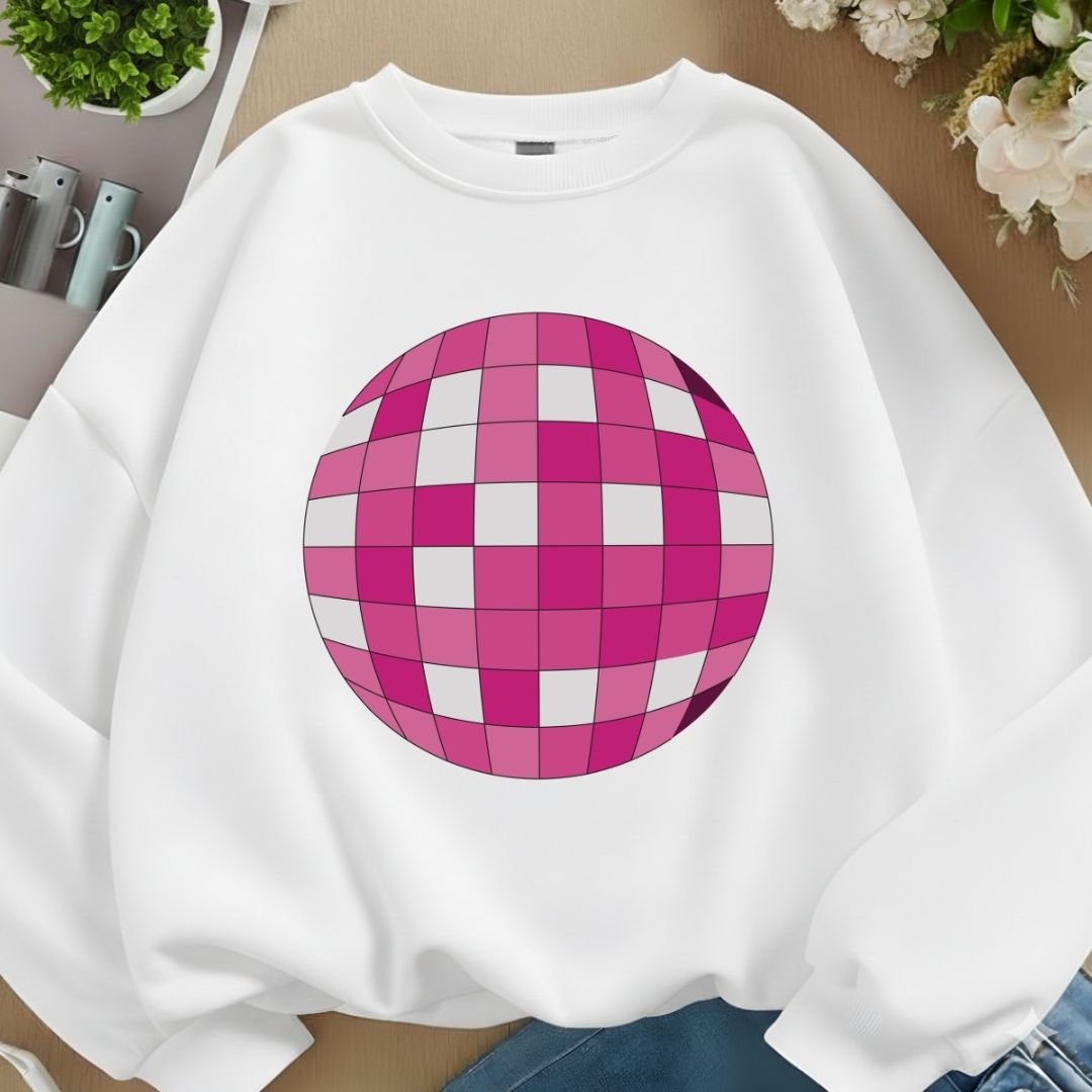 A white crewneck sweatshirt featuring a large pink and white checkered disco ball graphic on the front, laid flat on a wooden surface with decorative plants and flowers.