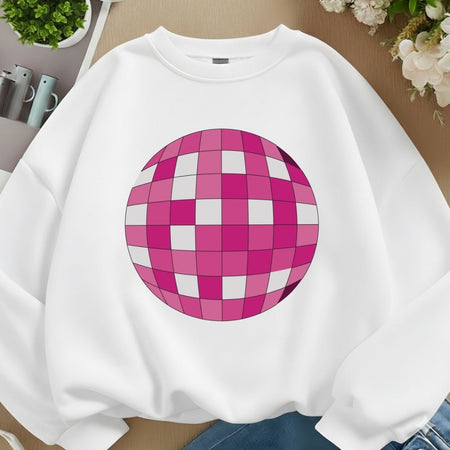 A white crewneck sweatshirt featuring a large pink and white checkered disco ball graphic on the front, laid flat on a wooden surface with decorative plants and flowers.