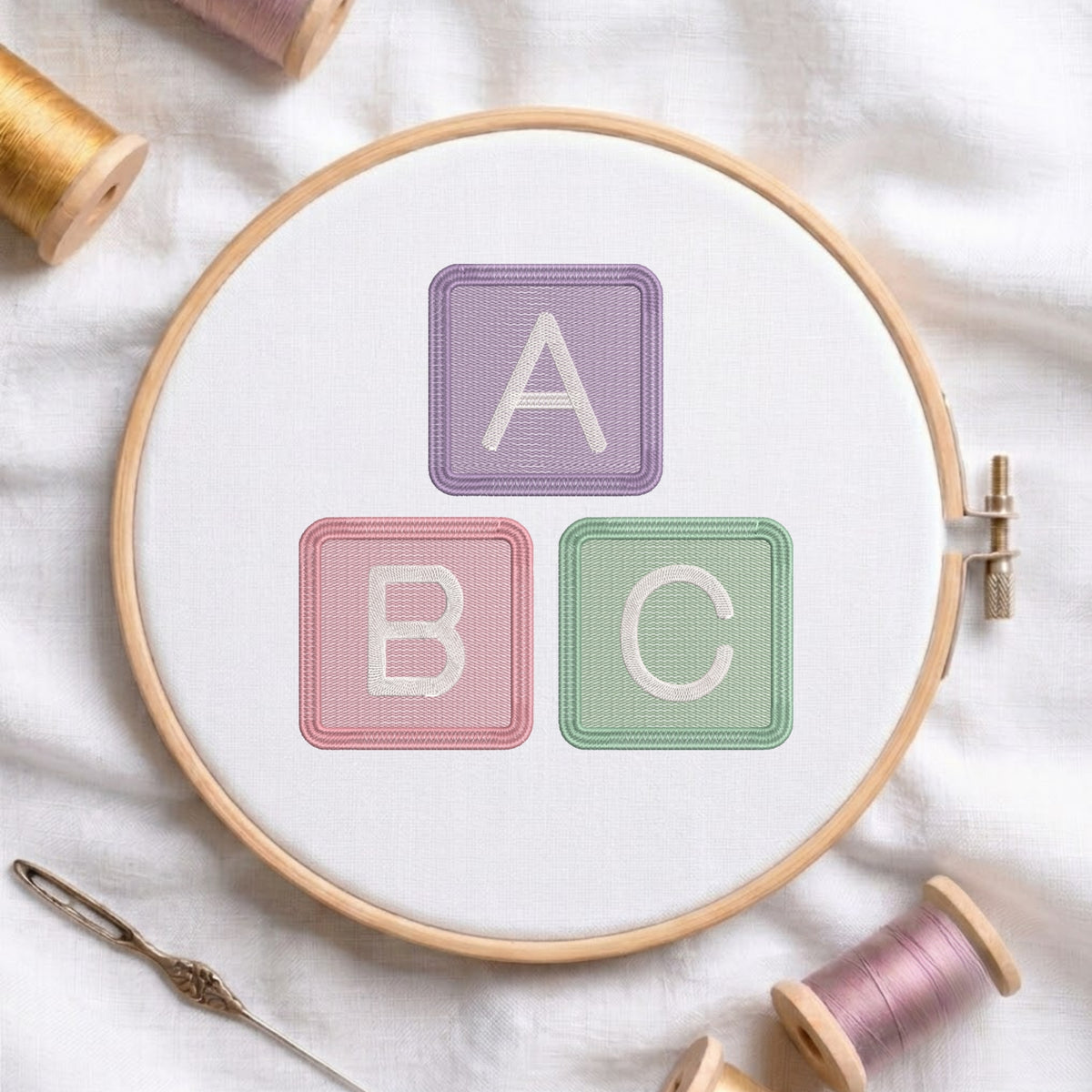 An embroidered alphabet blocks design spelling ABC in pastel purple, pink, and green stitched inside a wooden hoop on white fabric with sewing threads.