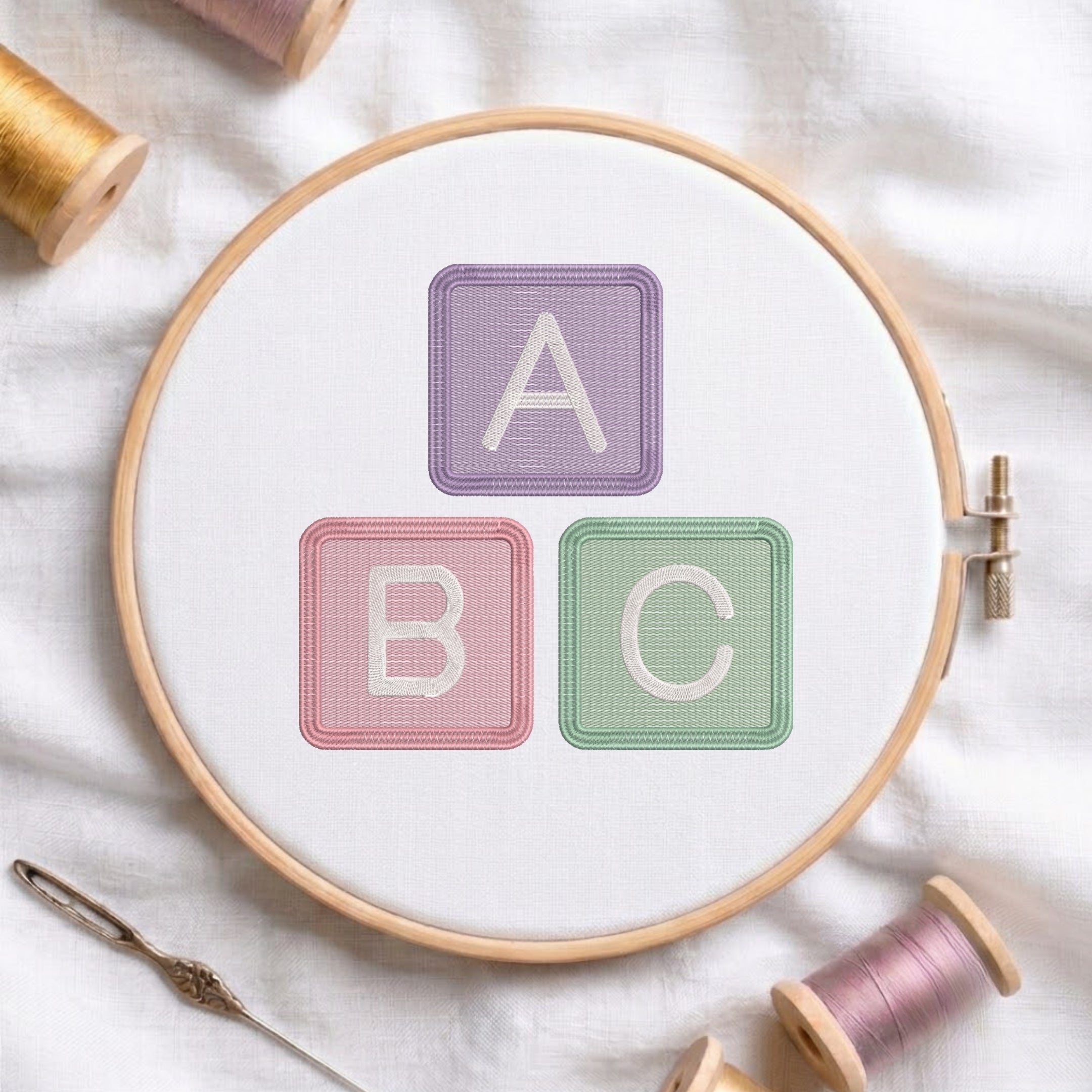 An embroidered alphabet blocks design spelling ABC in pastel purple, pink, and green stitched inside a wooden hoop on white fabric with sewing threads.