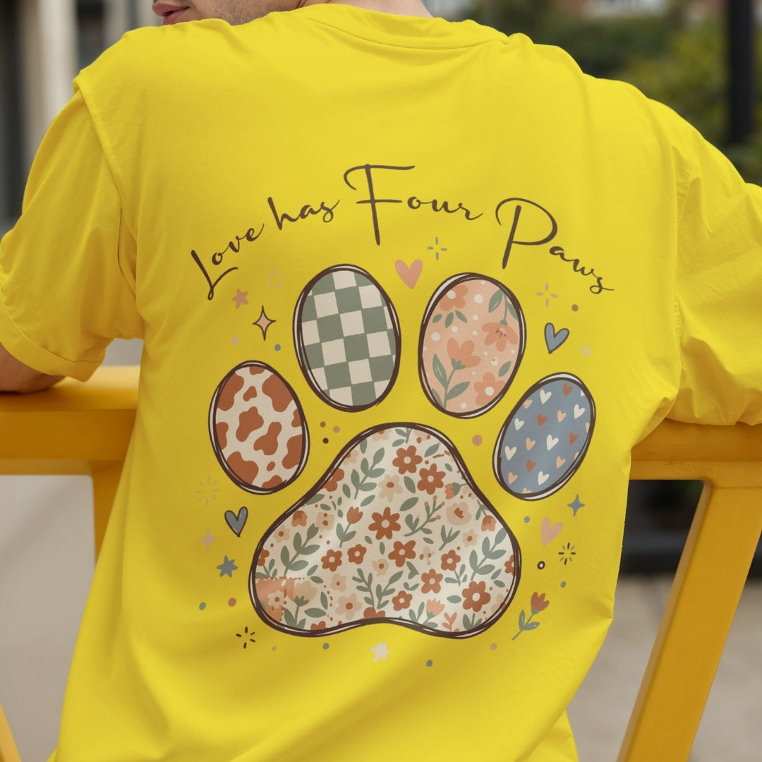 A bright yellow t-shirt worn by a person, showing the back with a large graphic of a paw print and the text 'Love has Four Paws' in a floral and patterned design.