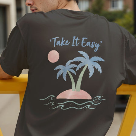 A man wearing a dark charcoal gray t-shirt, viewed from behind. The back of the shirt features a graphic print of a small tropical island with two blue palm trees, a pink sun, and light blue waves, with the text 'Take It Easy' written above.