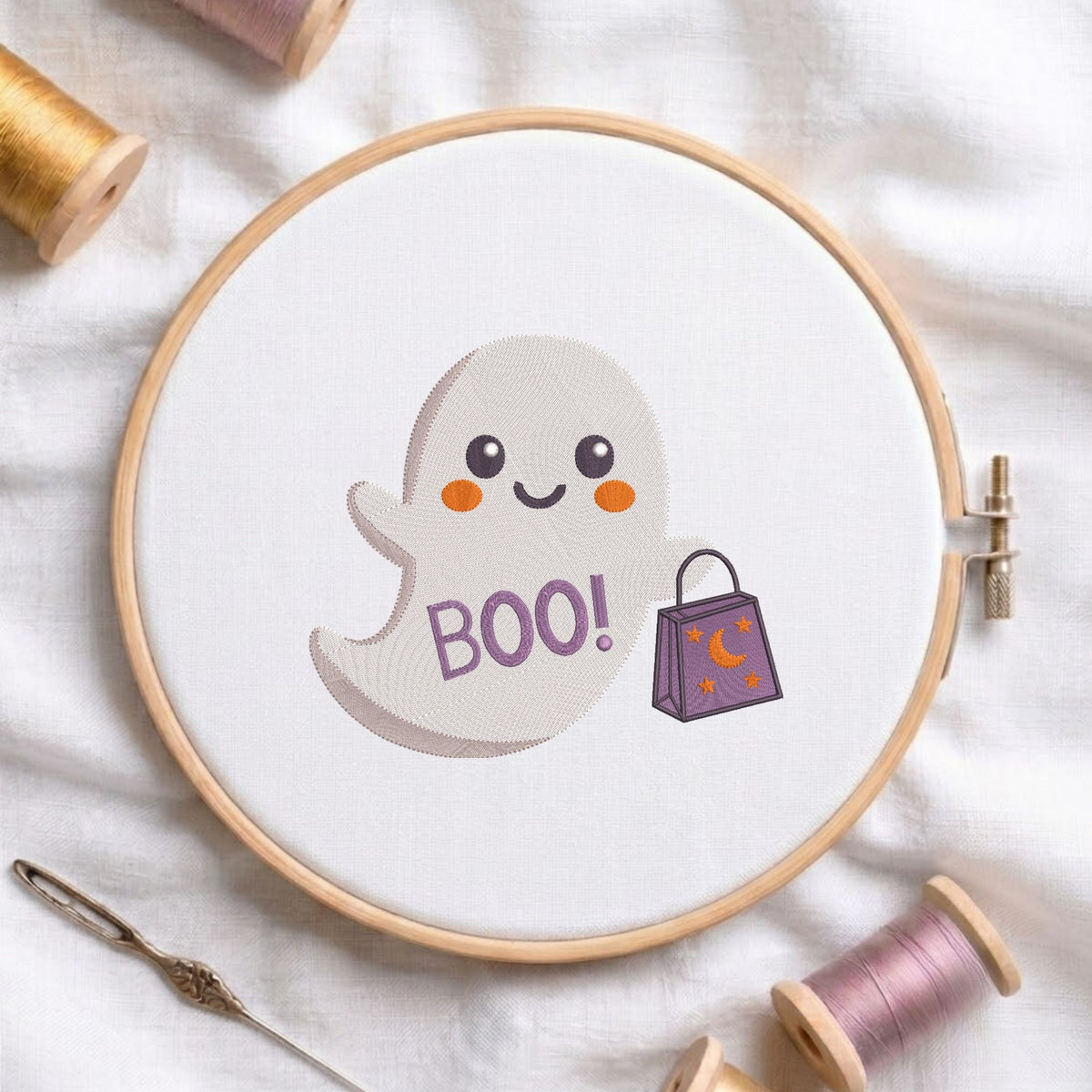 Cute ghost holding a "BOO!" shopping bag machine embroidery design displayed in a wooden hoop on white fabric with thread spools and a needle