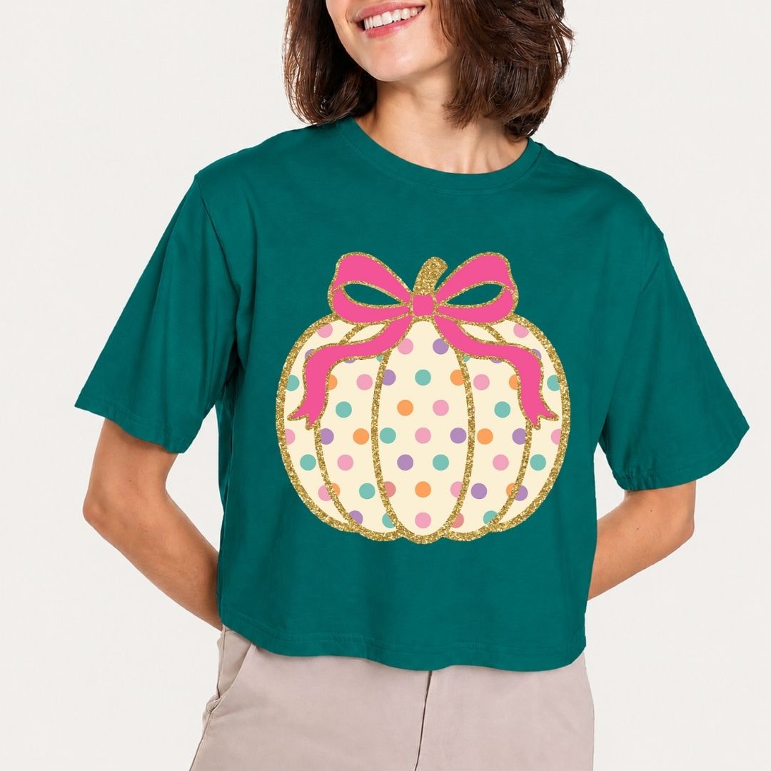 A woman wearing a dark teal short-sleeve crop top with a large graphic of a glittery gold pumpkin decorated with pink, purple, and orange polka dots and a pink bow, paired with light beige pants.