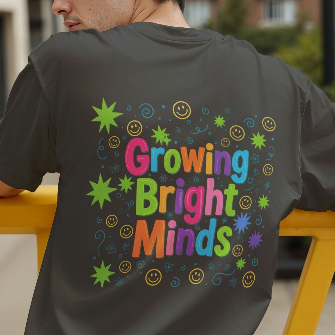 A man wearing a dark gray t-shirt, viewed from behind, showing a colorful graphic on the back that reads 'Growing Bright Minds' in rainbow letters surrounded by smiley faces, stars, and swirls.