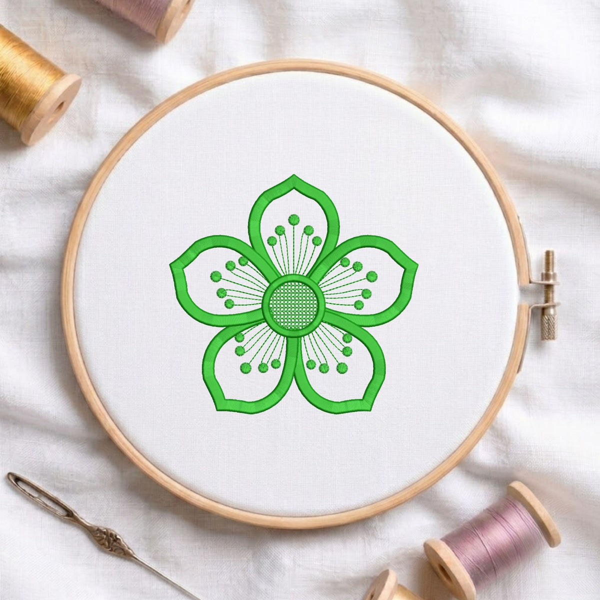 A wooden embroidery hoop holding white fabric with a bright green floral design stitched in the center. A needle and spools of thread are arranged around it on a white fabric background.