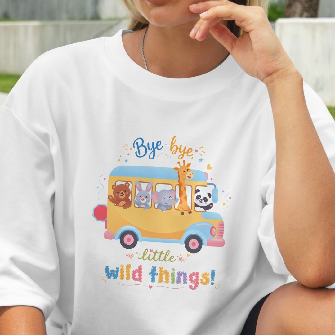 Person wearing a white t-shirt with a colorful graphic of a school bus and animals, with text 'Bye bye little wild things!'.