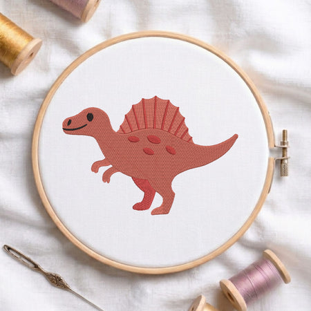 A wooden embroidery hoop holding white fabric with a stitched cartoon dinosaur in terracotta red. The hoop is styled on a white fabric background with spools of thread and a needle.