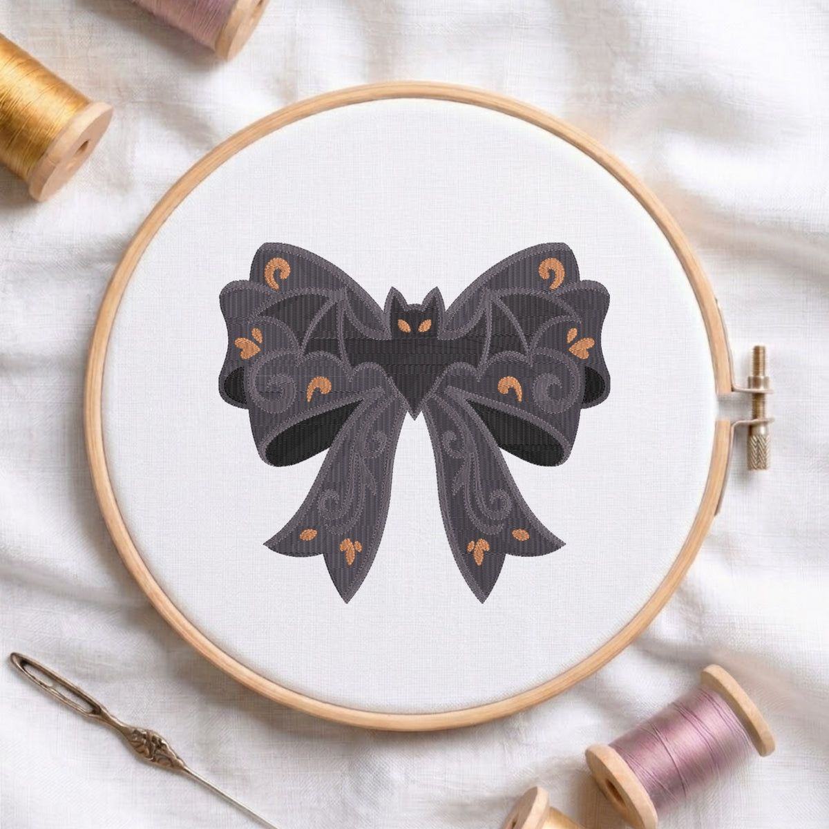 A white fabric stretched in a wooden embroidery hoop, featuring an embroidered dark bow patterned with swirling details, golden scrollwork, and a centered bat.c
