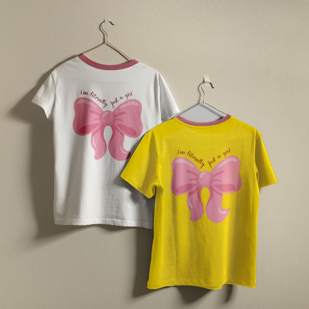 White and yellow t-shirts hanging on a wall, both showcasing the pink bow design and quote printed on the back.
