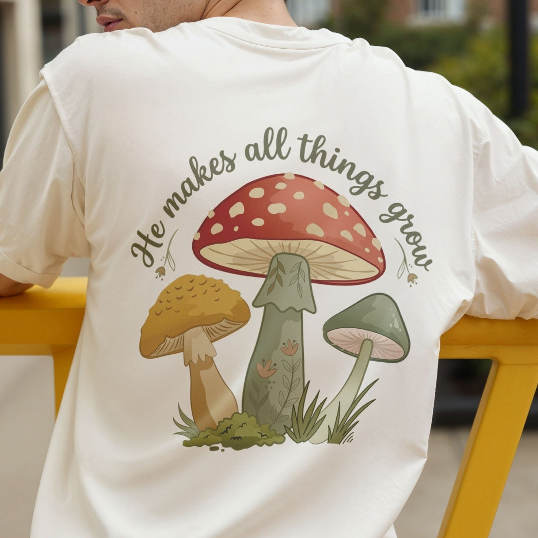 A person wearing a white graphic T-shirt, shown from the back. The shirt features a colorful illustration of three mushrooms and the printed phrase 'He makes all things grow' in dark gray script.
